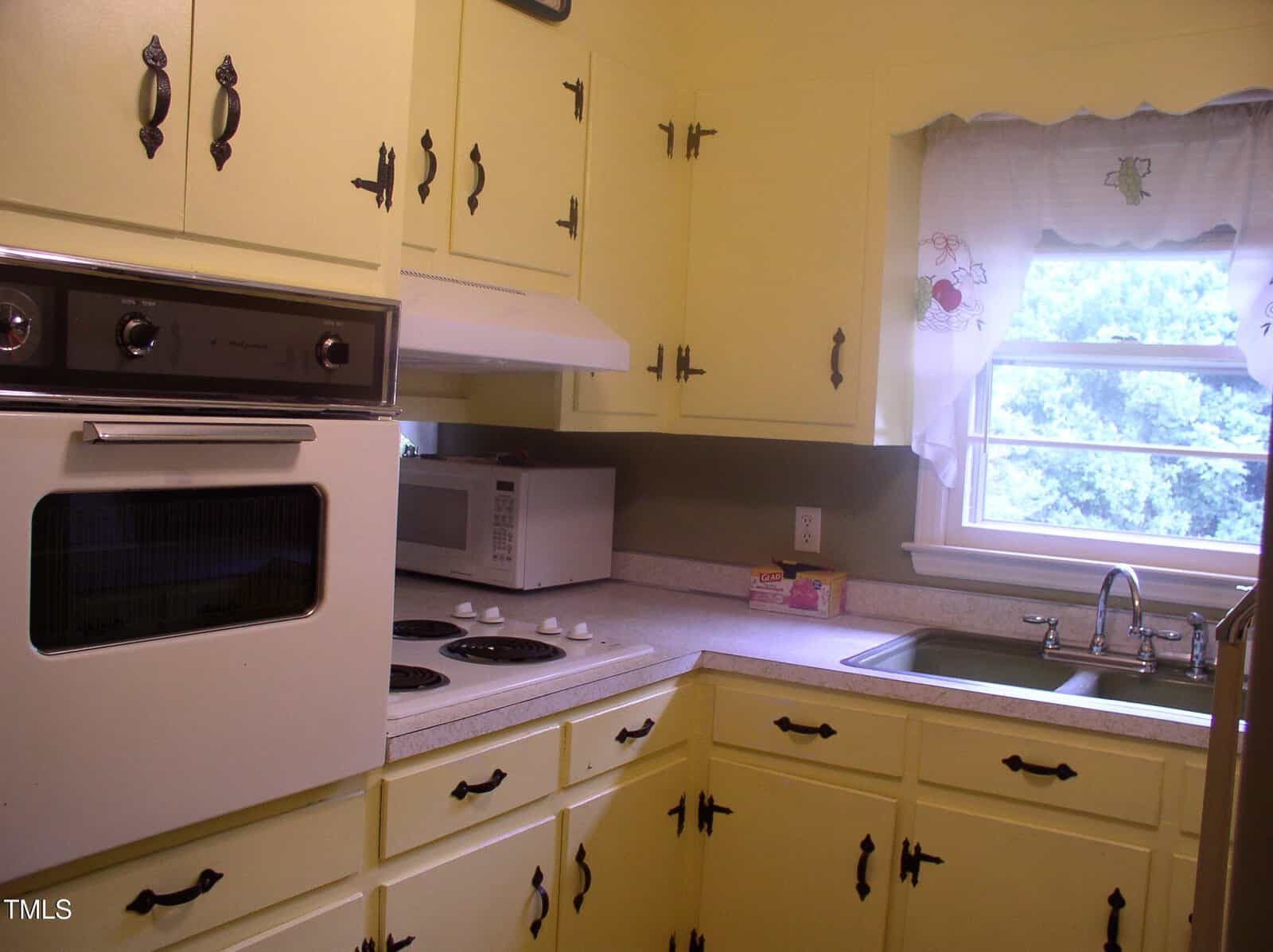 A kitchen with yellow cabinets, a wall oven, electric stove, microwave, sink under a window with white curtains, and a counter with a small box near the sink. Trees are visible outside the window.