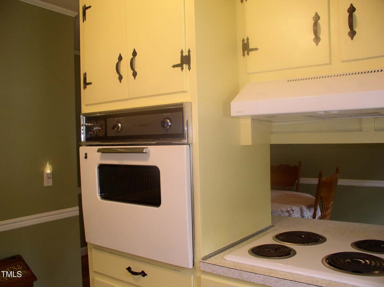 A vintage kitchen with yellow cabinets, a built-in wall oven, and an electric stovetop with four coil burners. A dining area with a wooden chair and table is partially visible in the background.