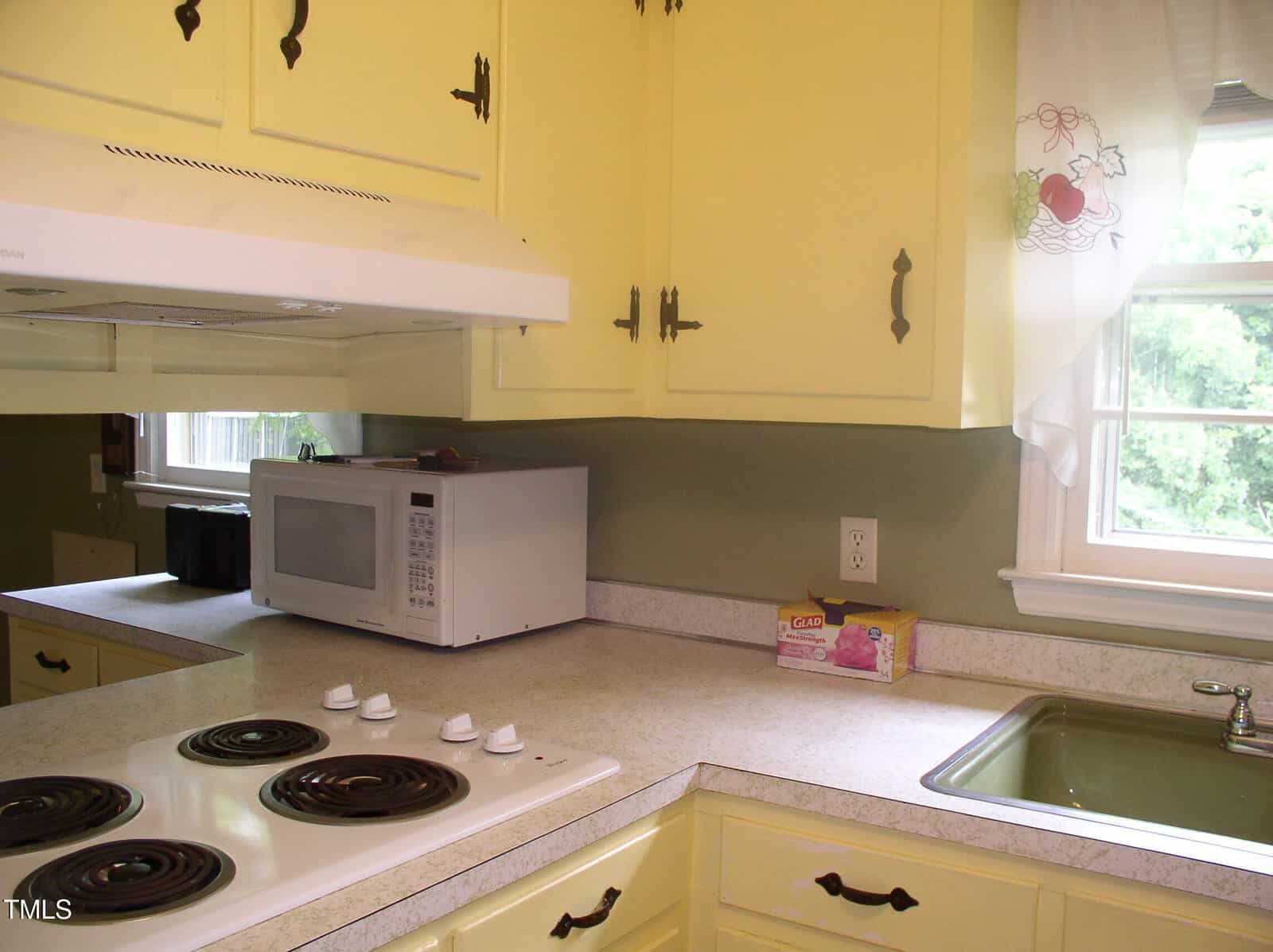 A kitchen with yellow cabinets, a white electric stovetop, a microwave, a box of Glad bags, a green sink, and a window with curtains showing trees outside.