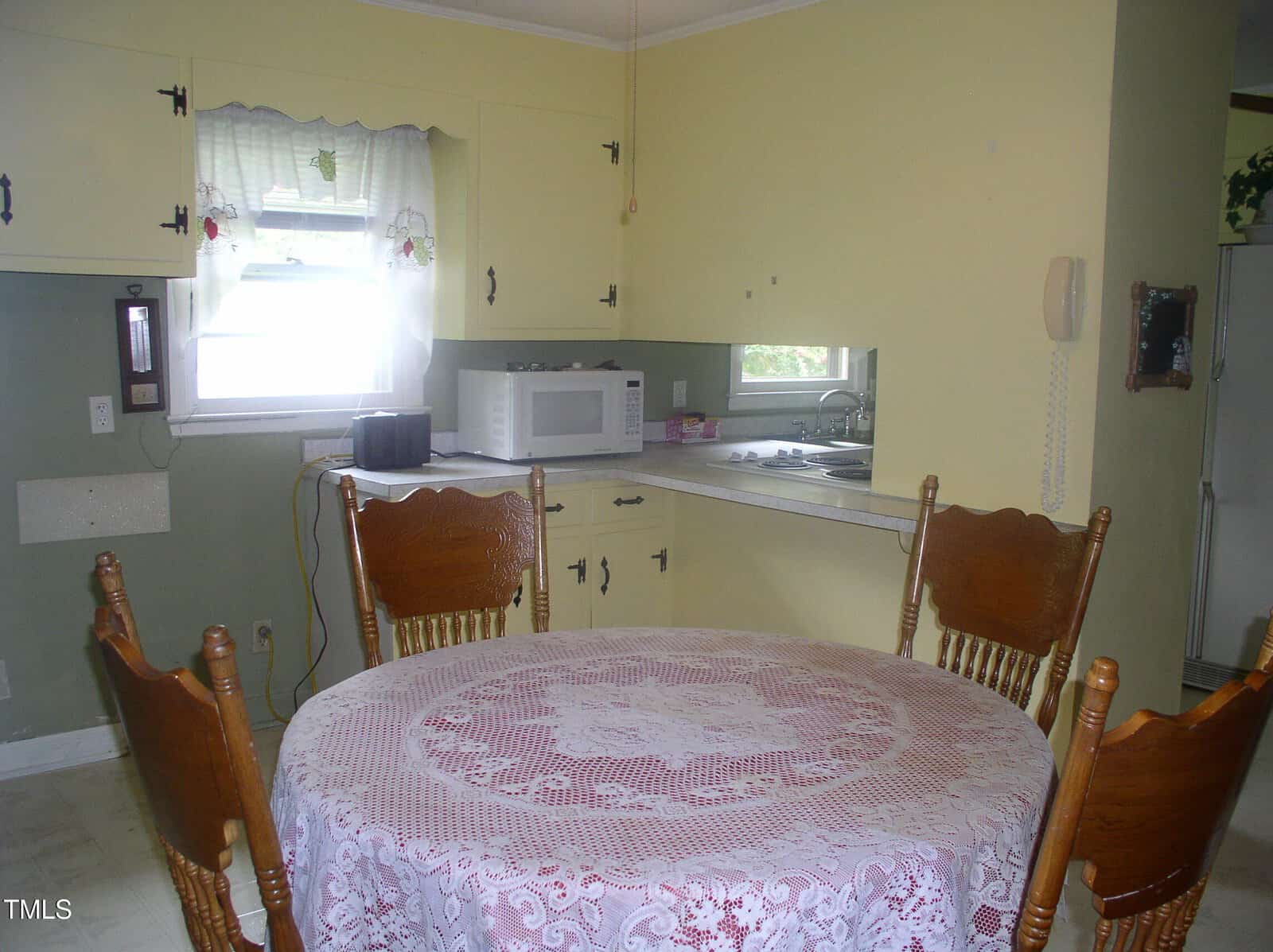 A small kitchen with a round table covered by a lace tablecloth, four wooden chairs, a microwave on the counter, a window with curtains, and a refrigerator in the corner.