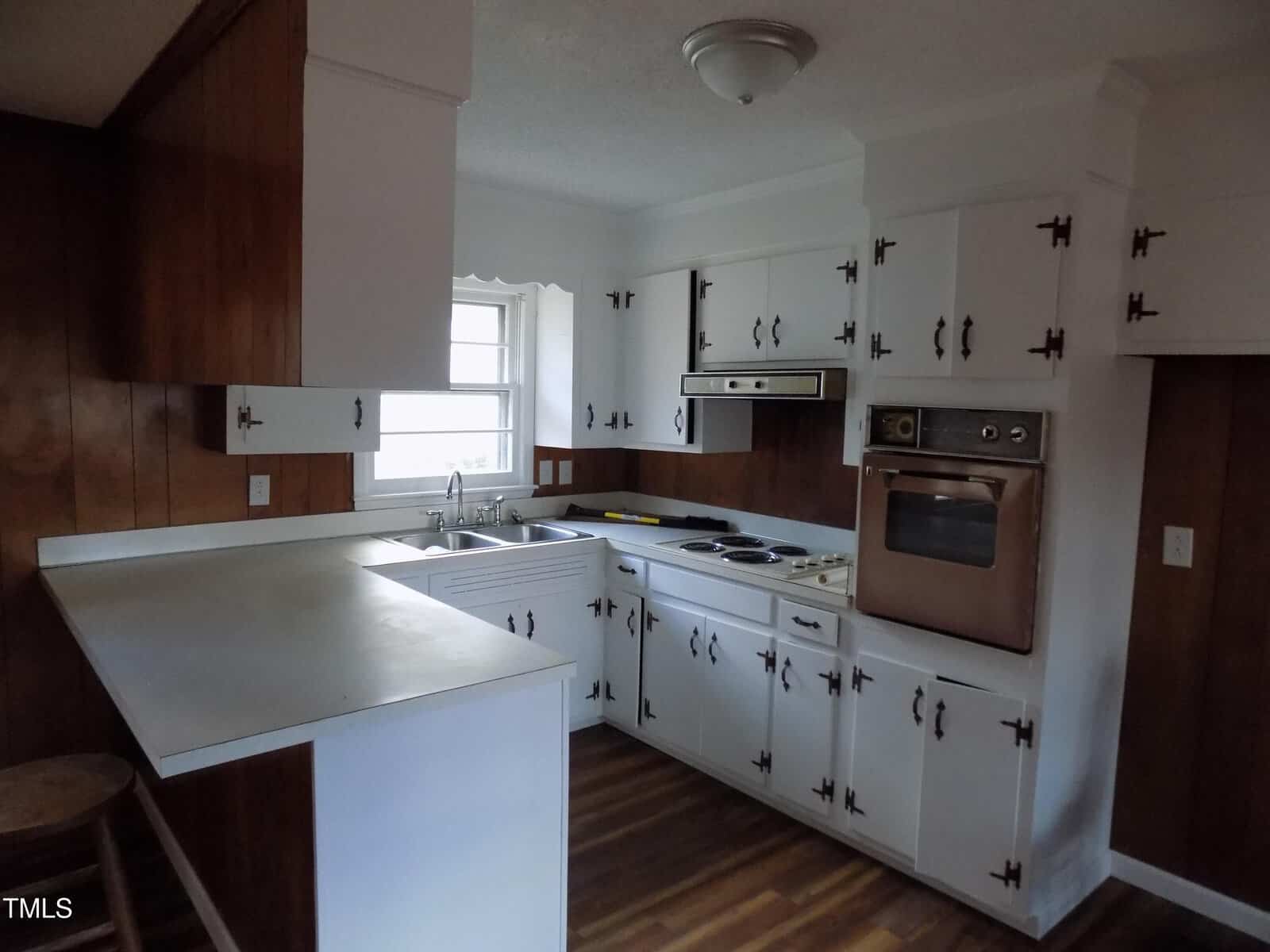 A small retro kitchen with white cabinets, wood-paneled walls, a built-in oven, electric stovetop, stainless steel sink, and a breakfast counter with stools. Natural light comes in through a window above the sink.