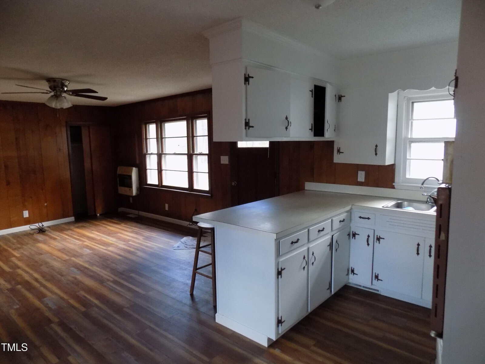 Open kitchen with white cabinets and countertop, next to a living area with wood panel walls, large windows, ceiling fan, and wood-style flooring. One upper cabinet door is open. Light enters from multiple windows.