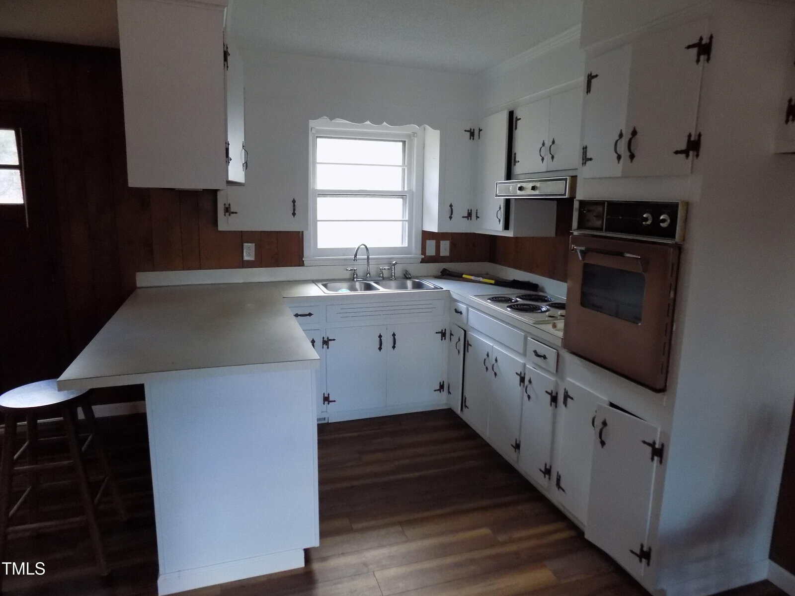 Small kitchen with white cabinets, wood accents, a window over the sink, an electric stove, a wall oven, and a breakfast bar with a wooden stool. The floor is wood-patterned, and appliances appear vintage.
