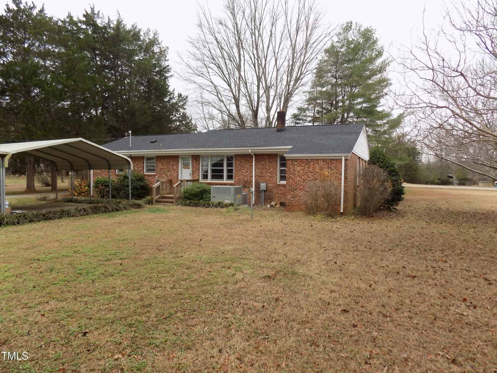 A single-story red brick house with a dark shingle roof sits on a grassy yard. There is a covered carport to the left, bushes near the foundation, and various trees surround the property.