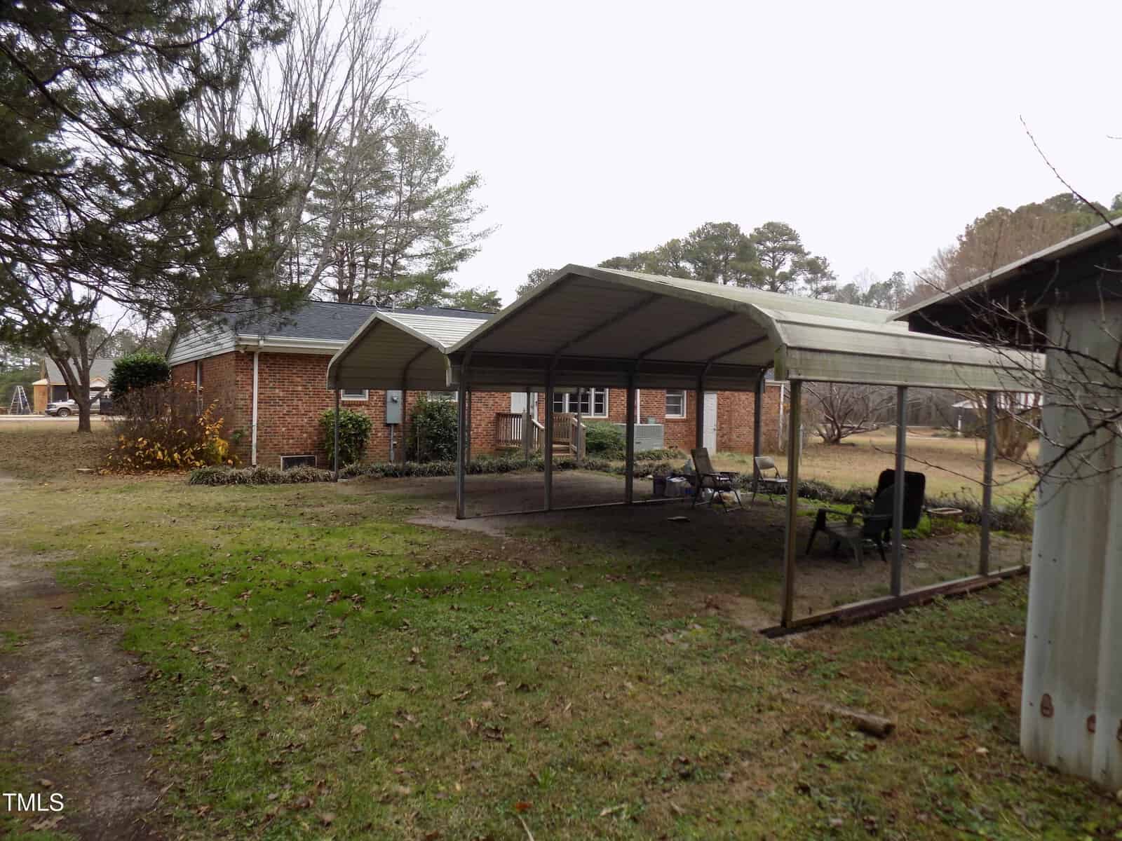 A brick house with a metal carport in front, set on a grassy yard with scattered trees and leaves. Lawn chairs and a small table are under the carport; an outbuilding is partially visible on the right.