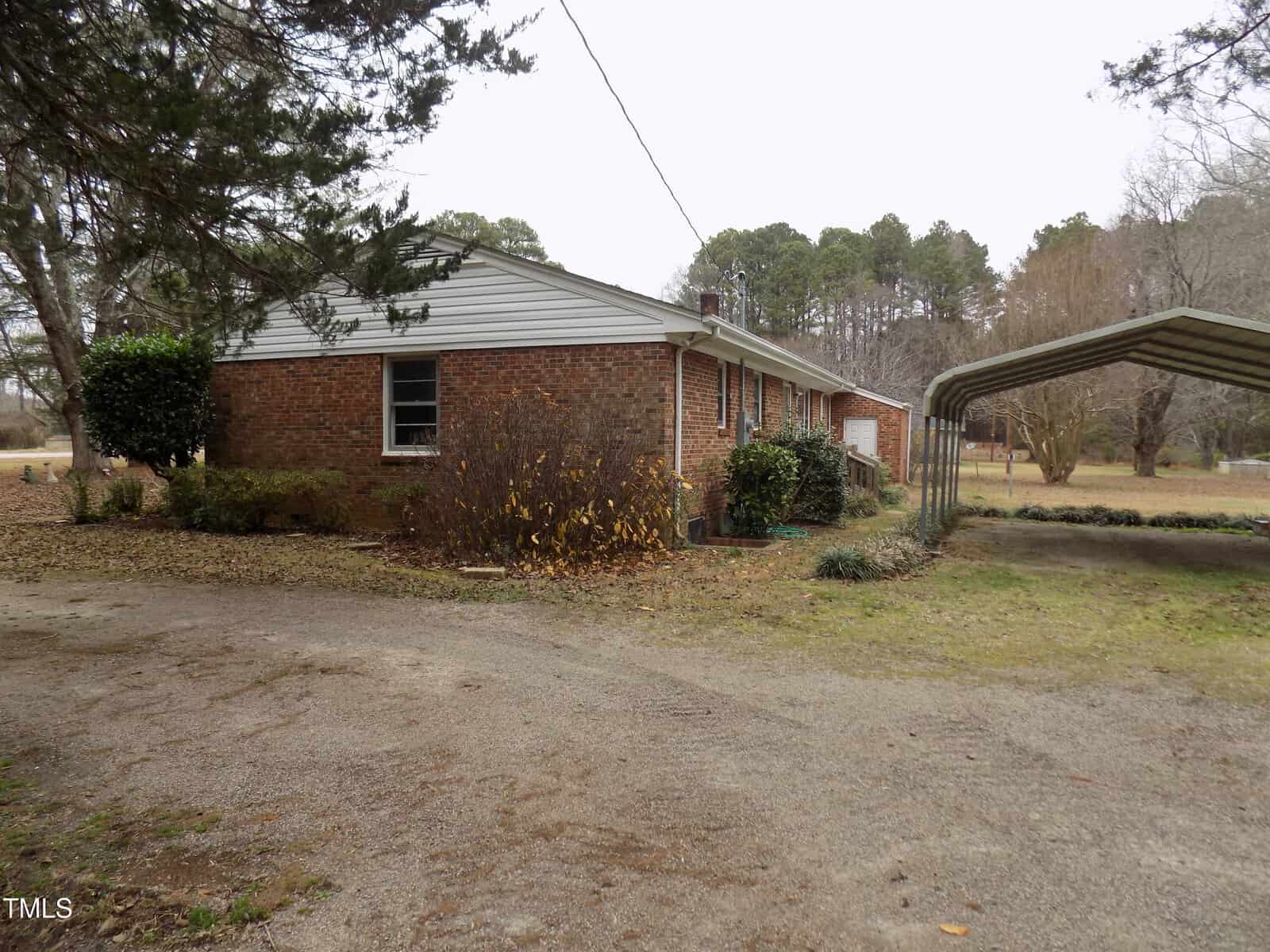 A single-story brick house with white siding under a cloudy sky, a gravel driveway, a metal carport, and trees in the background. Some leaves and shrubbery are visible near the house.