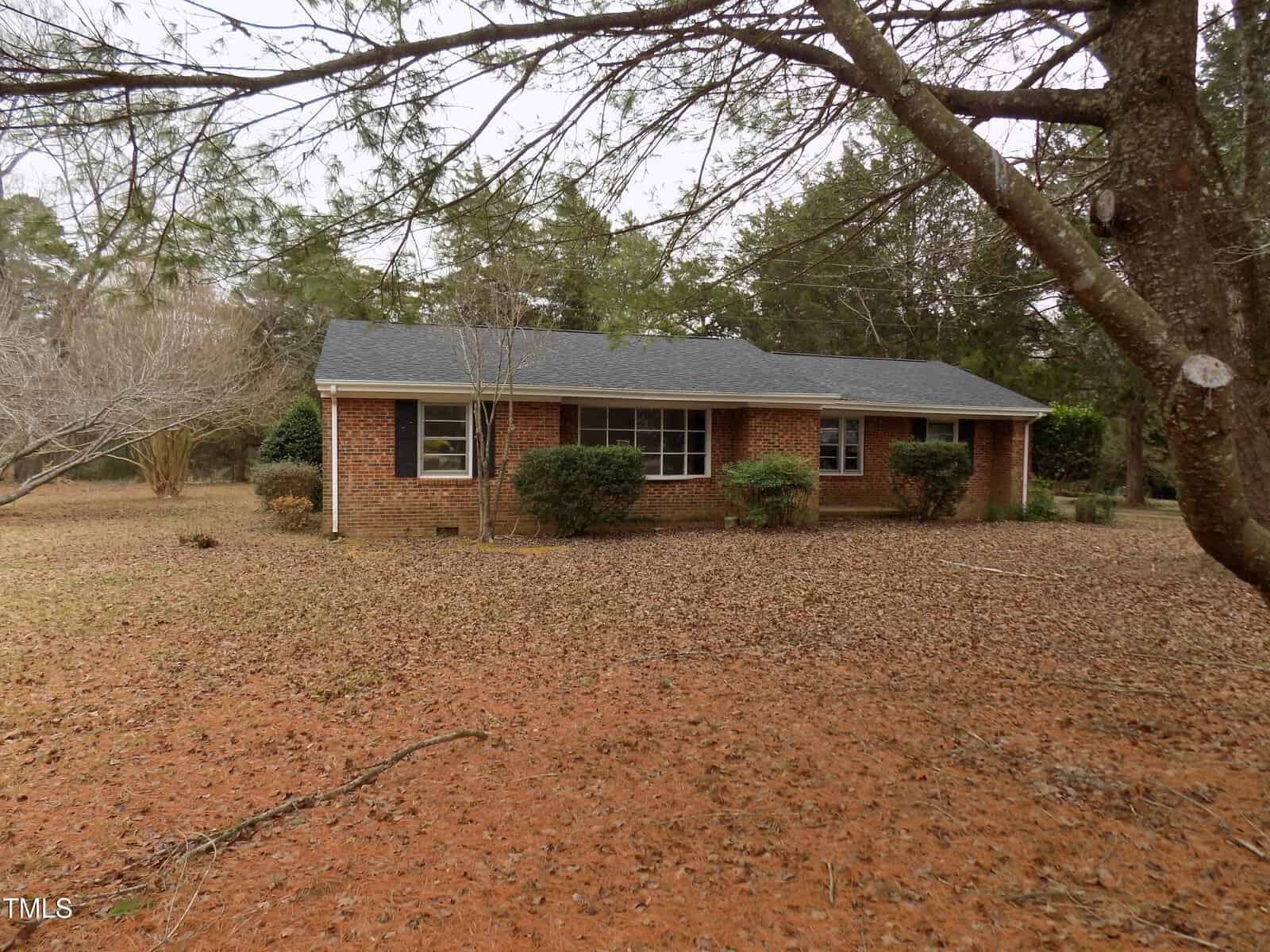 A single-story brick house with a dark shingled roof, large front windows, and a covered entryway, surrounded by trees and a yard covered in brown fallen leaves.