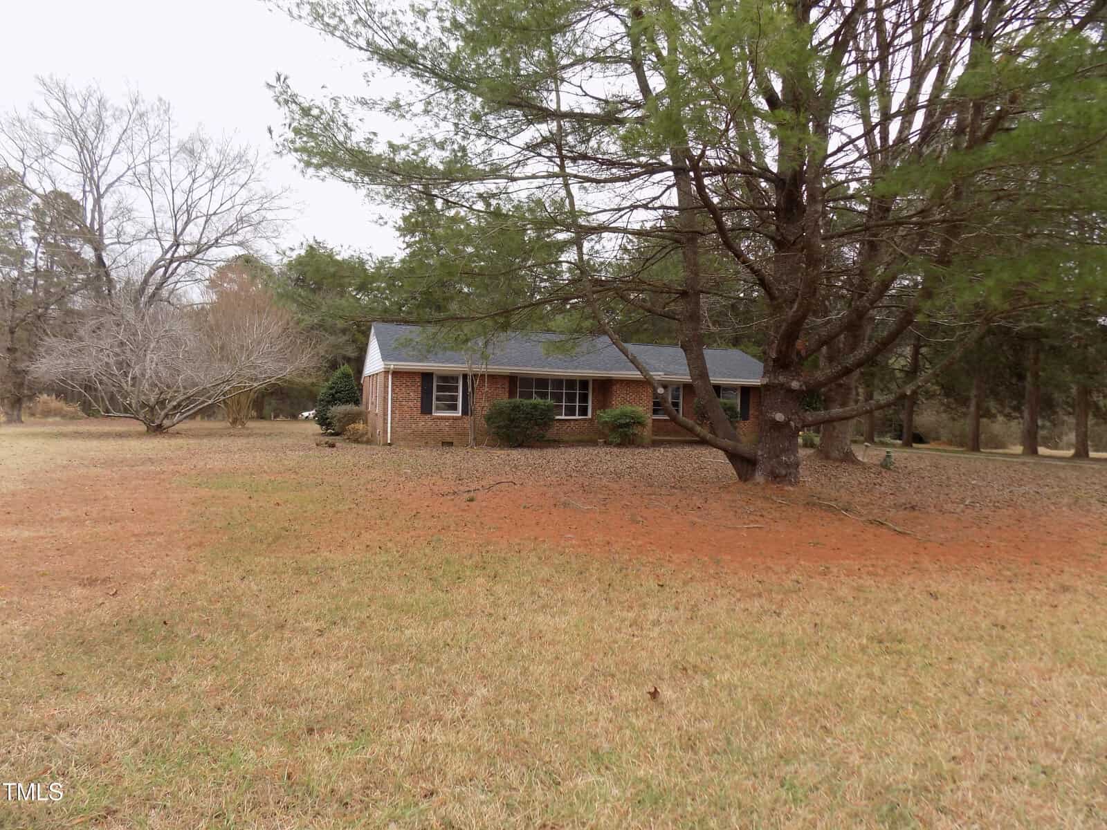 A one-story brick house with a dark roof sits on a grassy lawn, partially shaded by large pine trees. Leafless trees are visible in the background, and the sky appears overcast.