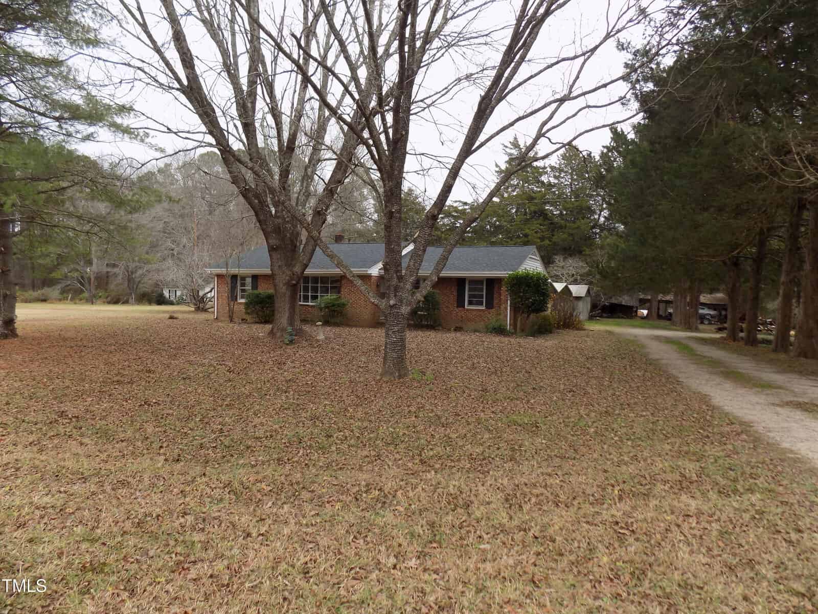 A single-story brick house with dark shingles is set back from a dirt driveway, surrounded by leafless trees and a large grassy yard under an overcast sky.