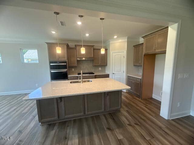 Modern kitchen with wood floors, a large center island with a sink, pendant lights, wood cabinets, a built-in oven, gas stove, and a pantry door. Walls are light-colored with natural and artificial lighting.