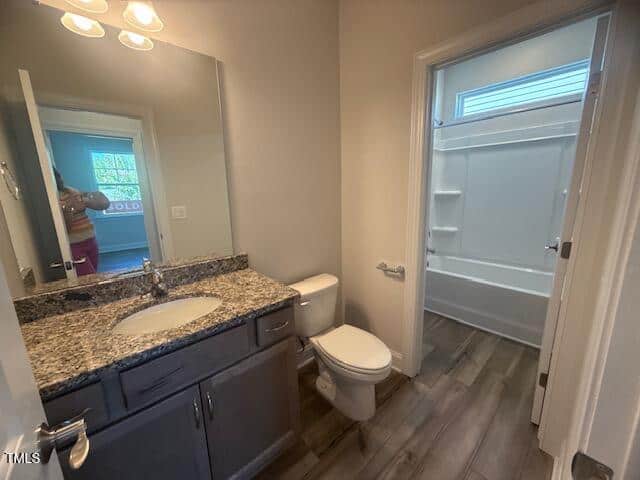A modern bathroom with a granite countertop vanity, sink, toilet, and a separate bathtub with built-in shelves. A person is partially visible in the mirror, and natural light comes through a window above the tub.