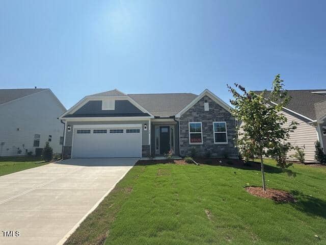 A single-story house with a stone and siding exterior, two-car garage, and a "sold" sign in front. The house has a well-kept lawn, driveway, and a newly planted tree, under a clear blue sky.