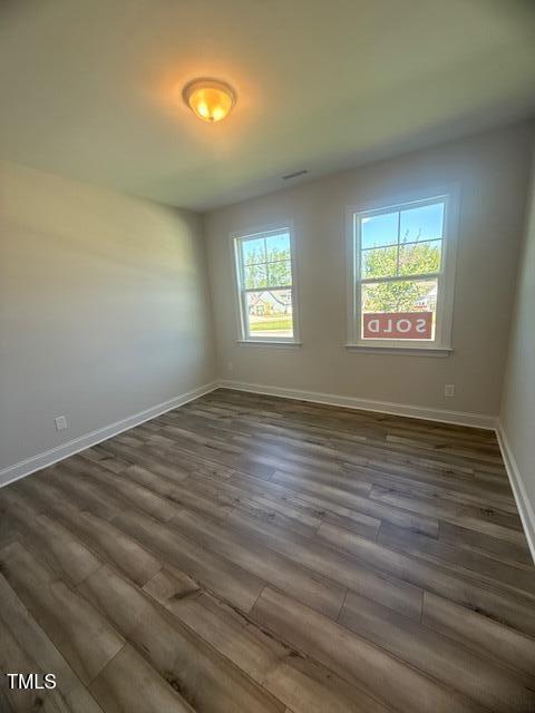 Empty room with light-colored walls and wood flooring, two large windows letting in natural light, and a "SOLD" sign visible outside. Ceiling light fixture is on.