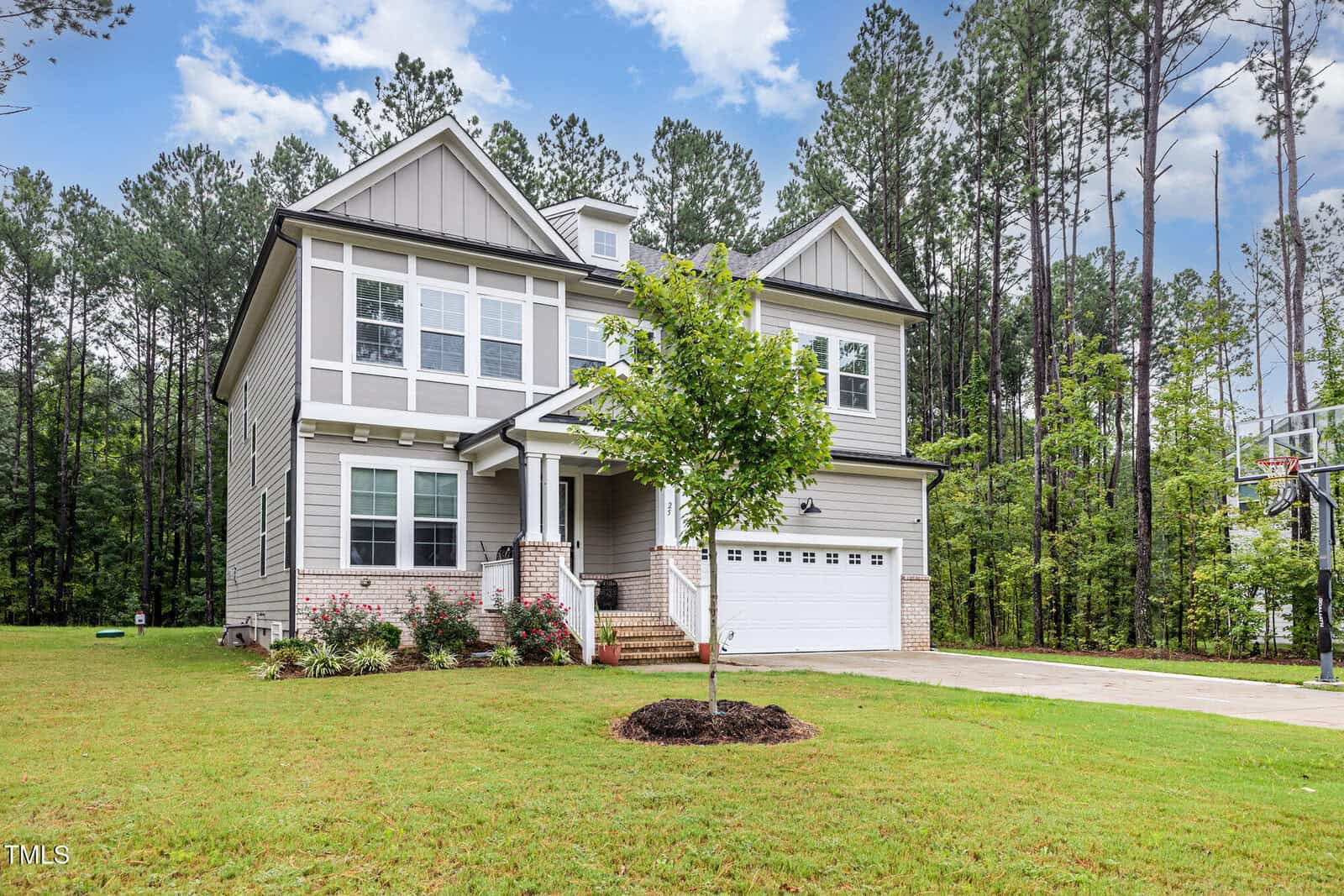 A modern two-story house with gray siding and white trim sits on a spacious lawn, surrounded by tall pine trees. A small tree is planted in the front yard and a basketball hoop is visible by the driveway.