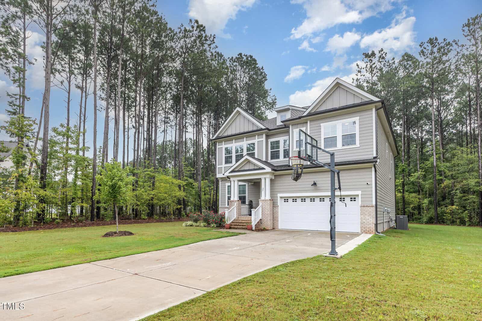 A two-story modern house with gray siding and white trim, a double garage, front porch, and a basketball hoop in the driveway. The home sits beside a lawn and is surrounded by tall pine trees.