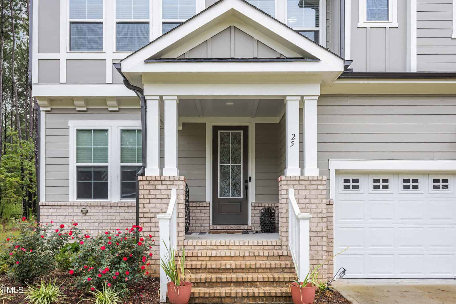 A modern house exterior with gray siding, white trim, a covered front porch with brick steps, a glass front door, potted plants, and a blooming rose bush next to a white garage door.