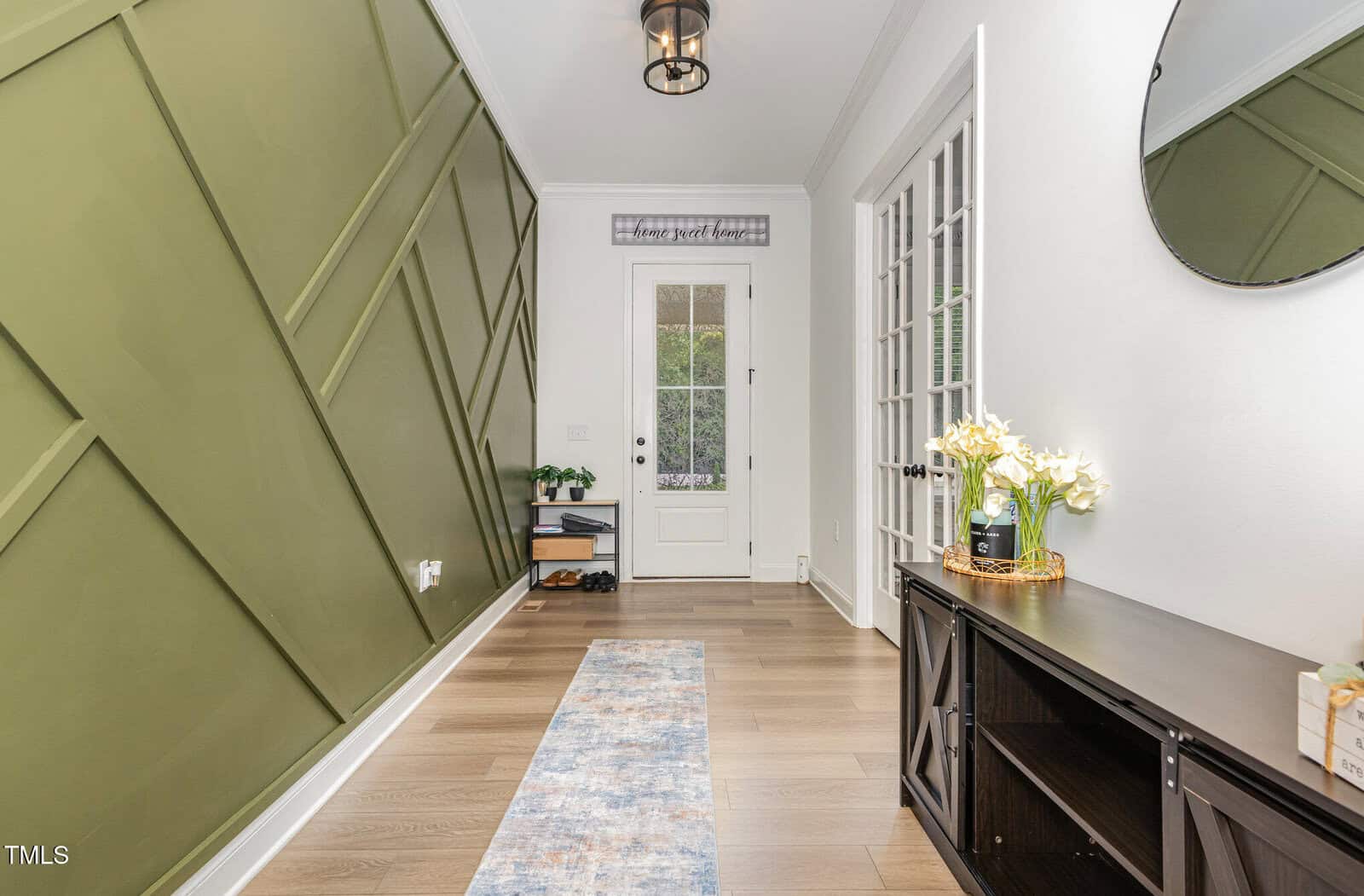 A modern entryway with light wood floors, a green accent wall with geometric patterns, a white front door with glass panes, a round mirror, a dark cabinet with flowers, and a patterned rug.