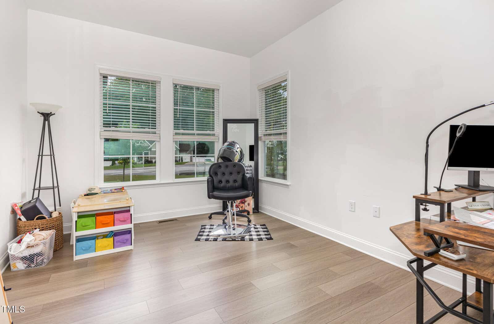A bright room with a salon chair on a checkered mat by a mirror, colorful storage bins, a basket, and a computer desk with a monitor. Large windows let in natural light and overlook a green outdoor scene.