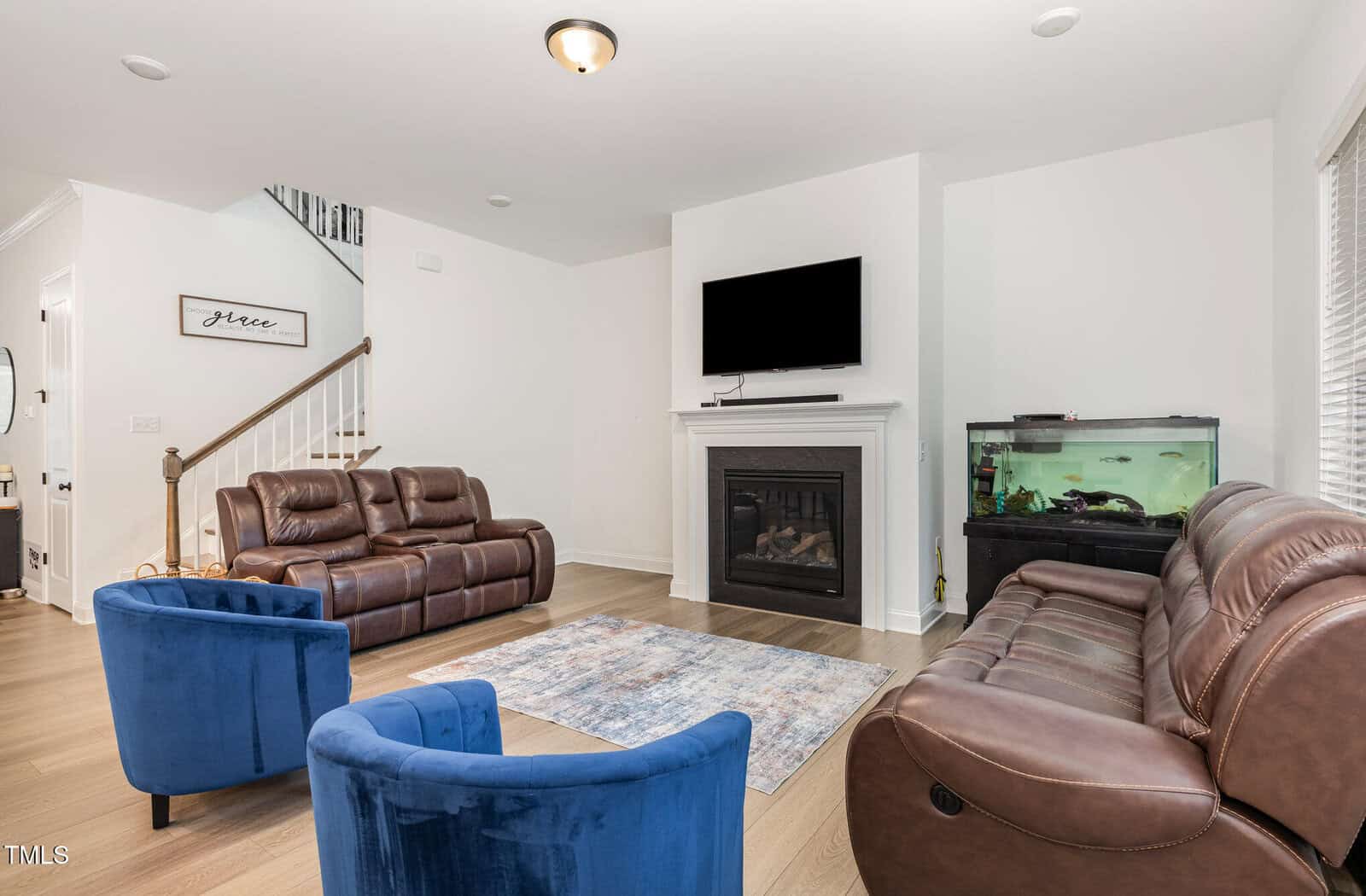 Modern living room with brown leather sofas, two blue armchairs, a fireplace with a TV above it, a fish tank to the right, wooden floor, and a staircase with white railing in the background.