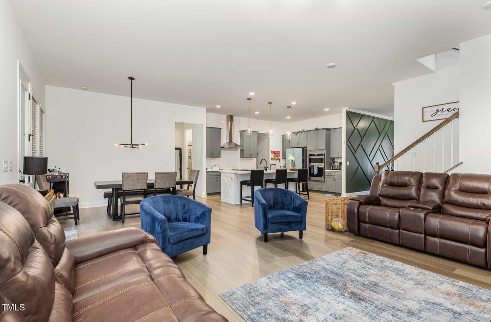 Open-concept living space with brown leather sofas, two blue armchairs, a rug, and a dining area. The modern kitchen has white cabinets, an island, pendant lights, and stainless steel appliances.