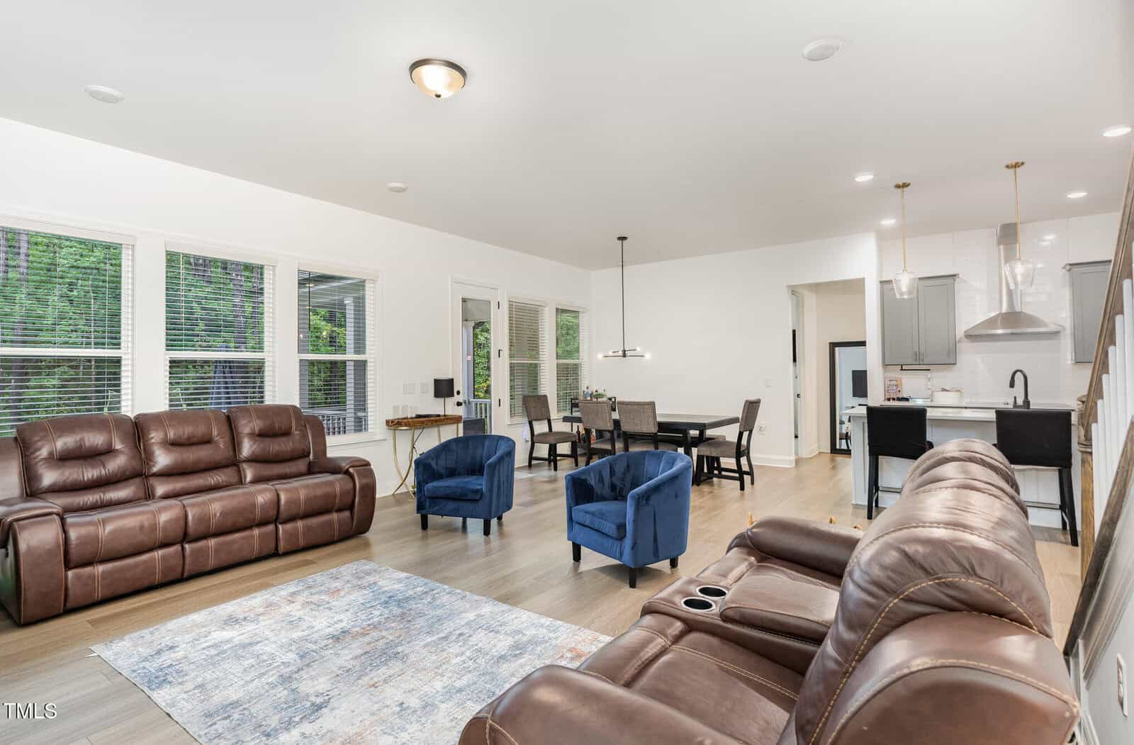Modern open-concept living area with brown leather sofas, two blue armchairs, a dining table, and a white kitchen. Large windows let in natural light, and light wood floors run throughout the space.