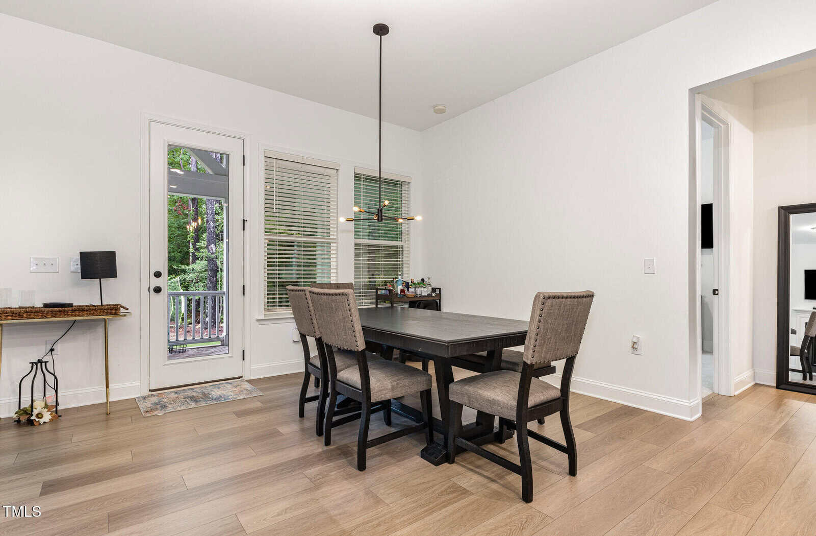 A modern dining area with a dark wood table, four upholstered chairs, a hanging light fixture, and large windows near a glass door leading outside. Light wood flooring and minimal decor complete the room.