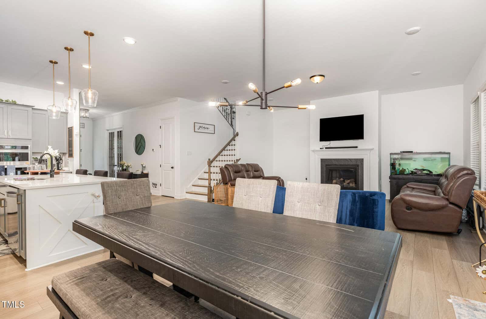 Modern open-concept living and dining area with a dark wood table, upholstered chairs, kitchen island with pendant lights, a staircase, brown recliners, a fireplace, mounted TV, and a fish tank.