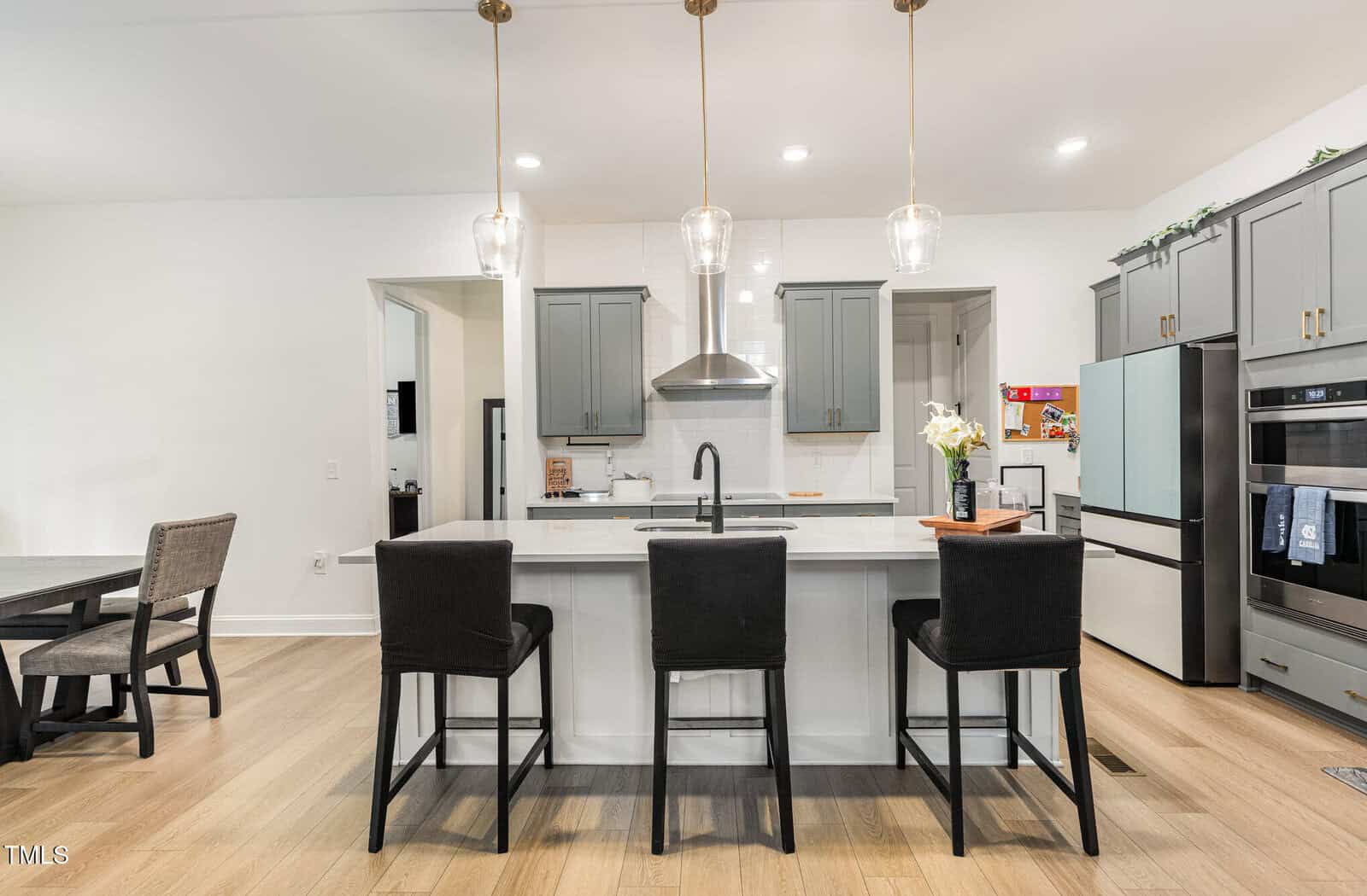 Modern kitchen with light wood flooring, gray cabinets, a white island with three black chairs, pendant lights, stainless steel appliances, and a built-in oven. A dining table is visible to the left.