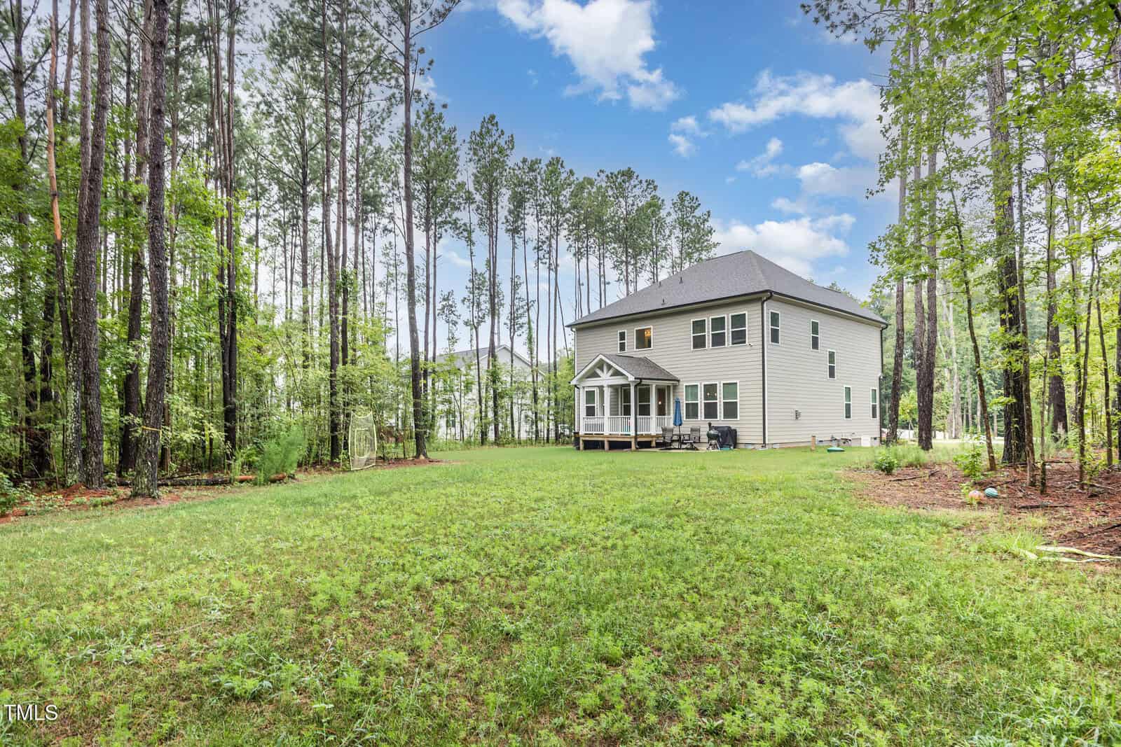 A two-story house with gray siding sits in a spacious grassy backyard surrounded by tall pine trees under a partly cloudy blue sky. A small covered porch is visible at the back of the house.
