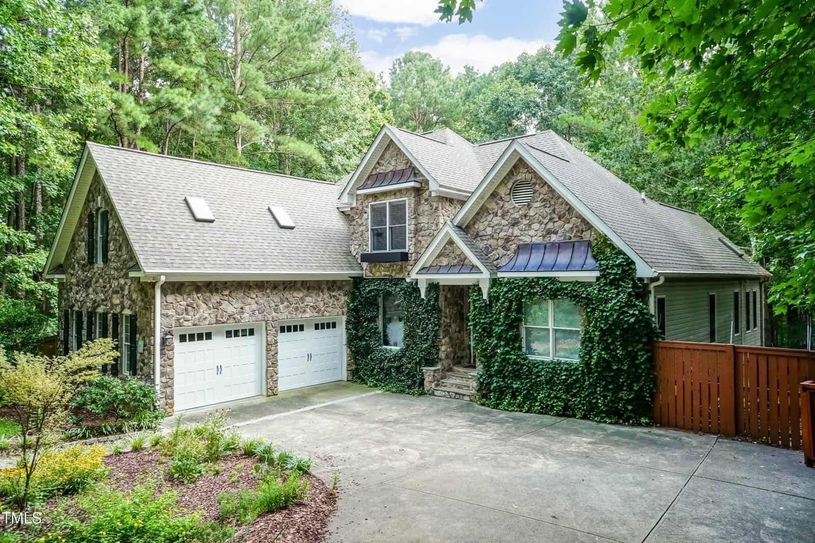 A two-story house with a stone exterior at 65 Hidden Cove Court, Youngsville, features ivy climbing the front, three white garage doors, and a paved driveway, all surrounded by trees and greenery.