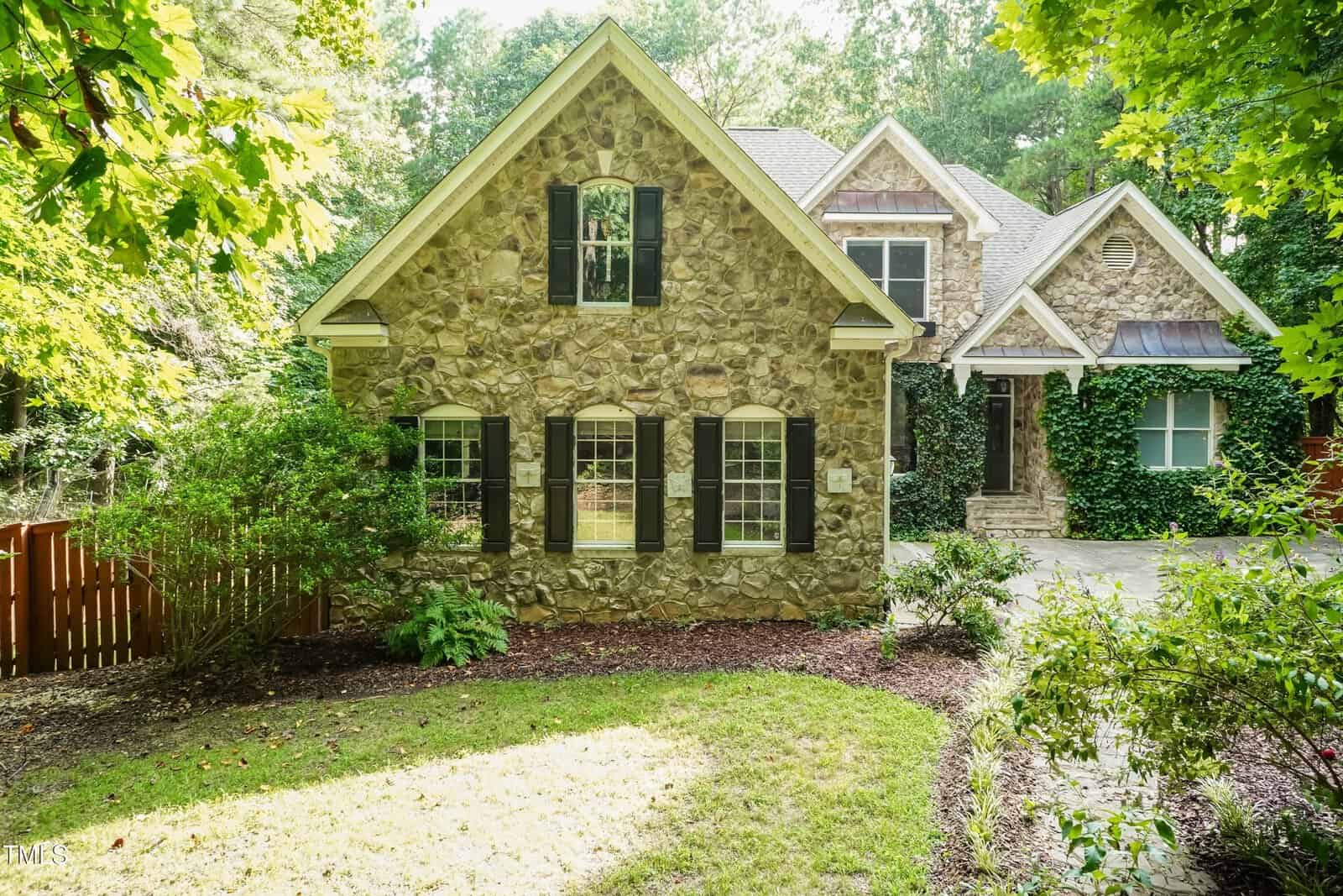 A two-story house with a stone exterior, black shutters, and multiple windows. The front yard has green shrubs, trees, and a wooden fence to the left, with a driveway on the right side.