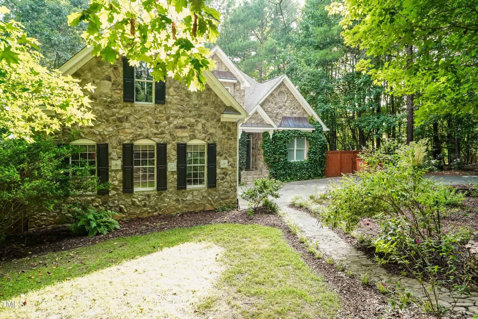 A stone house with multiple large windows, surrounded by trees and greenery, with a paved walkway leading to the entrance and a wooden fence in the background.