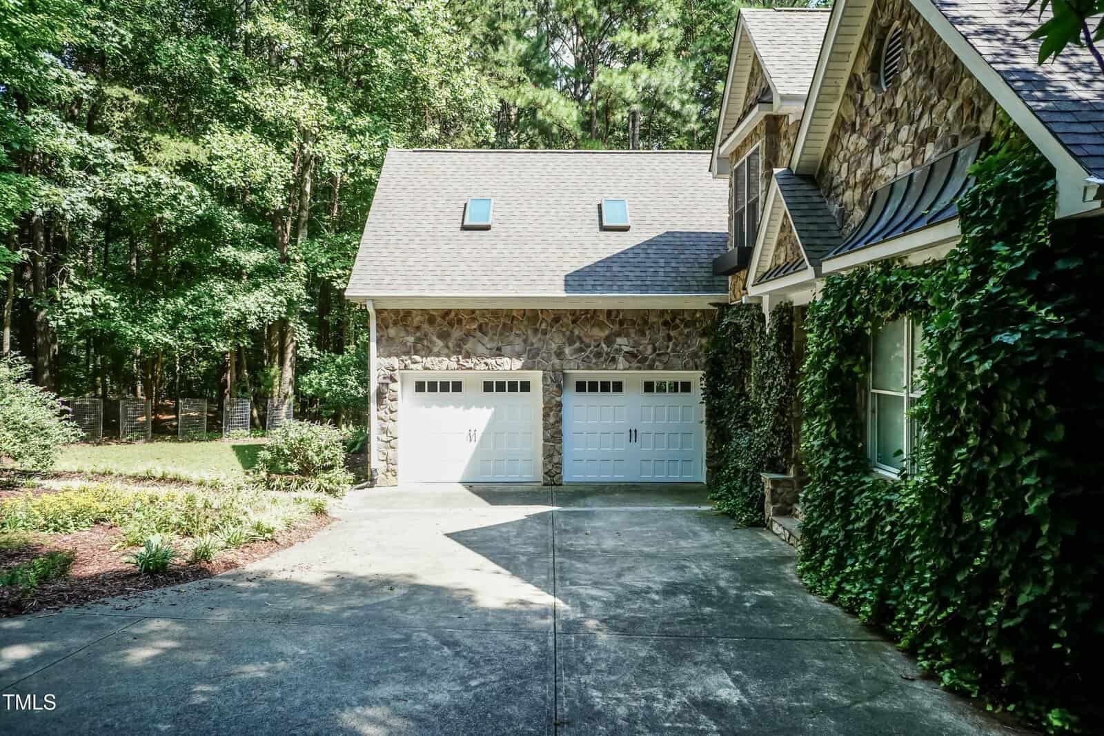 A two-car garage with white doors is attached to a house with stone siding and ivy climbing up the exterior walls. A concrete driveway and landscaped yard with trees are visible.