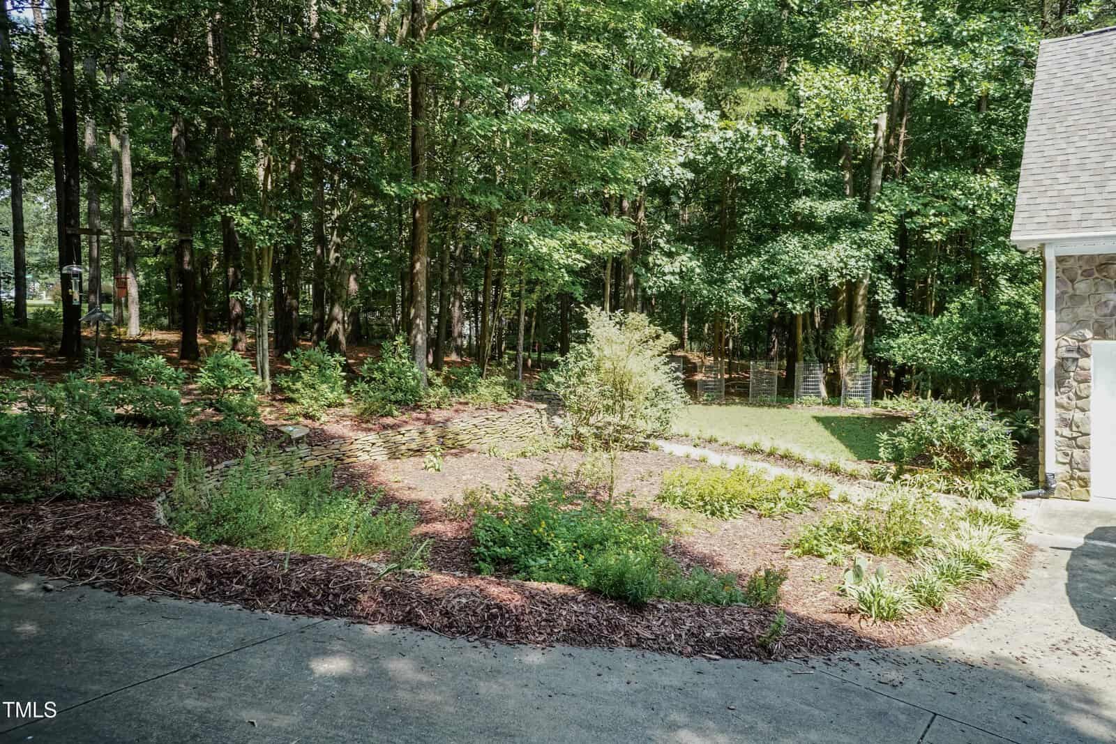 A landscaped garden bed with mulch and various small shrubs and plants sits next to a concrete driveway, bordered by tall trees in a wooded area.