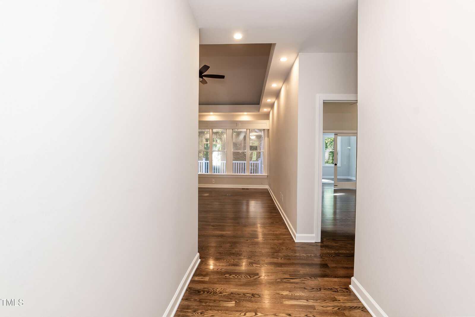 A hallway with hardwood floors leads to a living area with tray ceiling, recessed lighting, ceiling fan, and large windows showing a view of a porch and greenery outside.