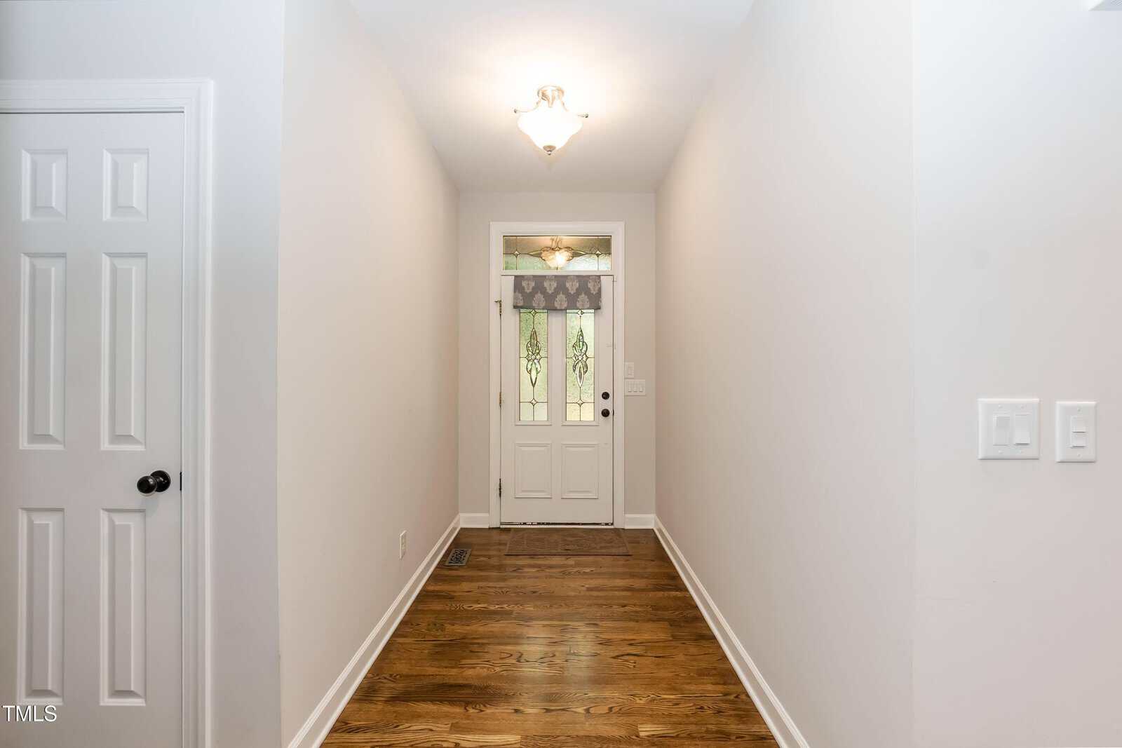 A hallway with hardwood floors, white walls, a light fixture on the ceiling, and a white front door with decorative glass and a valance. Two light switches and a vent are visible on the right wall and floor.