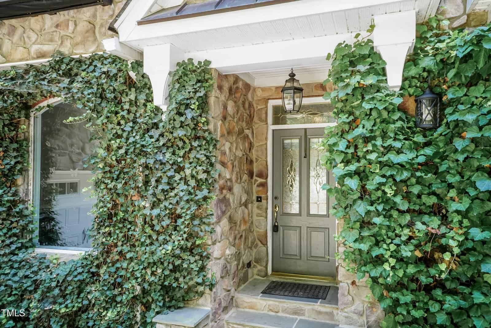 A stone house entryway with a gray front door, sidelight windows, a hanging lantern light fixture above, and green ivy climbing around the walls and window.