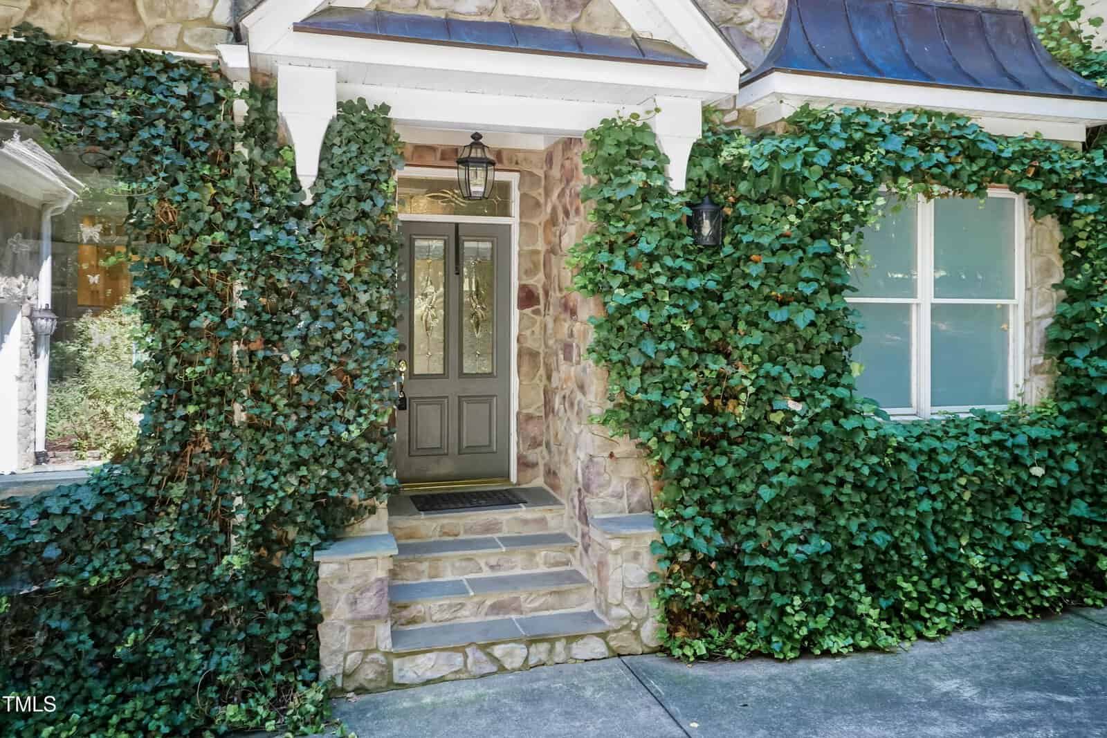 A stone house entrance with a gray door featuring decorative glass panels, two lantern-style wall lights, and ivy growing around the doorway and windows. Stone steps lead up to the front door.
