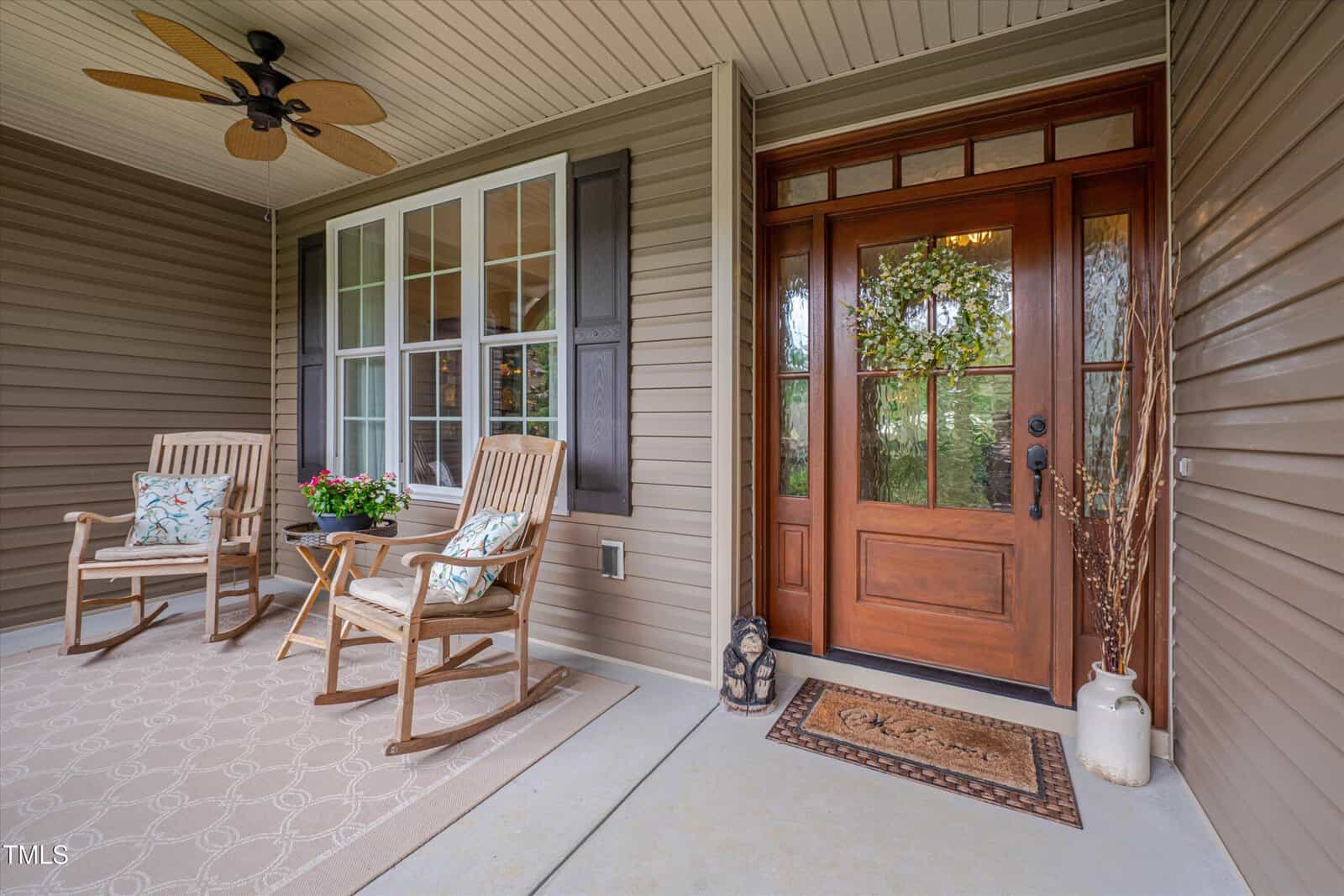 Covered porch with two rocking chairs, a small table with flowers, a ceiling fan, and a wooden front door with glass panels and a wreath. Welcome mat and decorative items are on the doorstep.