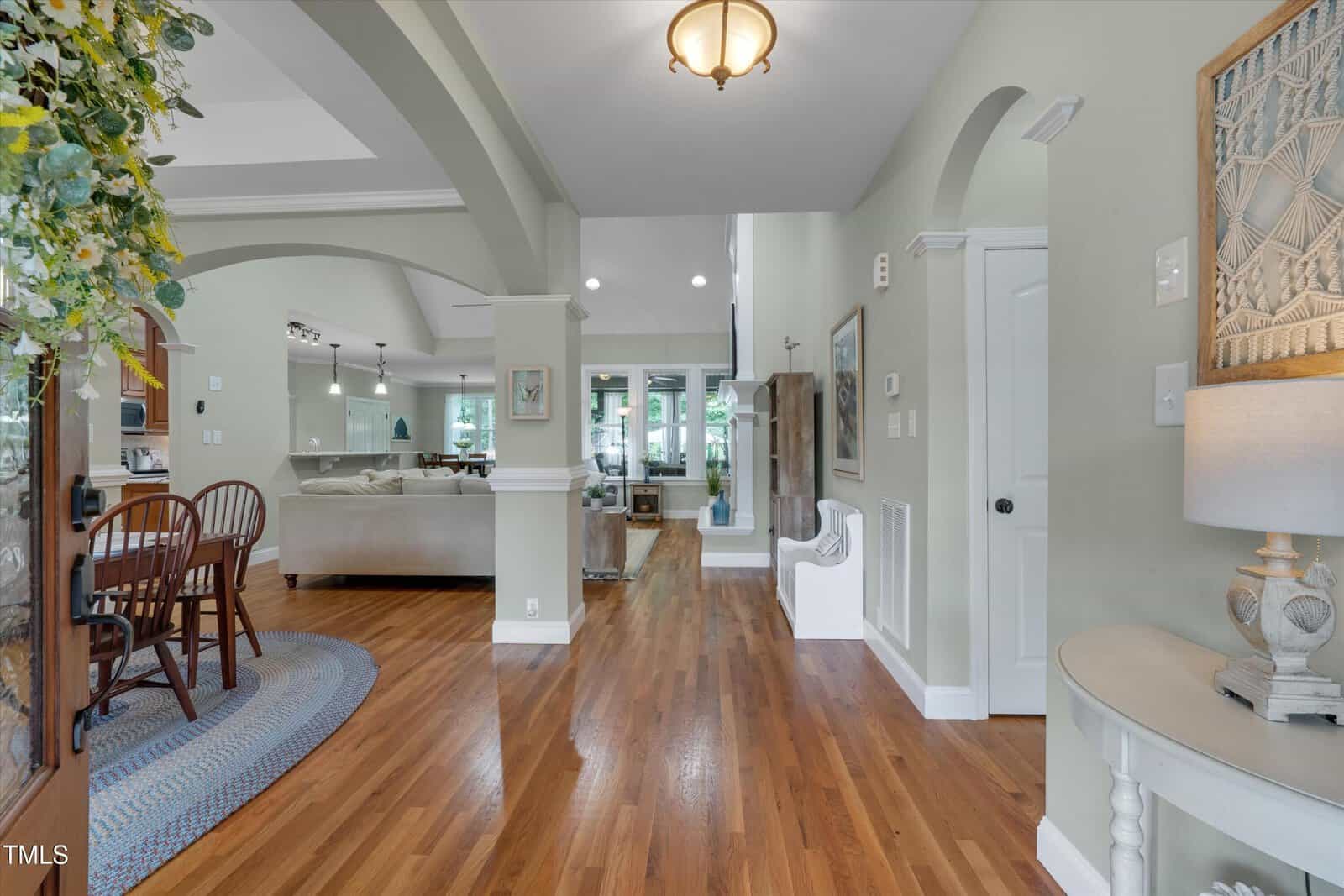 A bright entryway with hardwood floors leads to an open living and dining area. The space features neutral walls, recessed lighting, arched doorways, and large windows providing natural light.