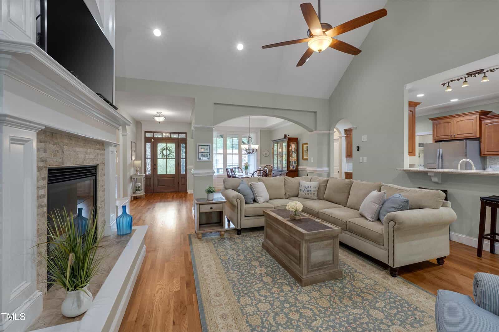 Spacious living room with beige sectional sofa, patterned rug, fireplace, ceiling fan, hardwood floors, and light green walls. Open layout leads to dining area and kitchen with wooden cabinetry.
