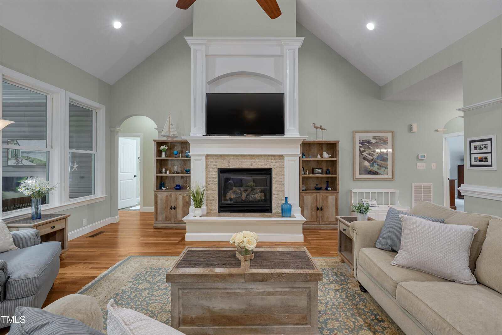 A cozy living room with a fireplace, TV above it, wooden shelves on both sides, beige sofas, a wooden coffee table with flowers, and large windows letting in natural light.