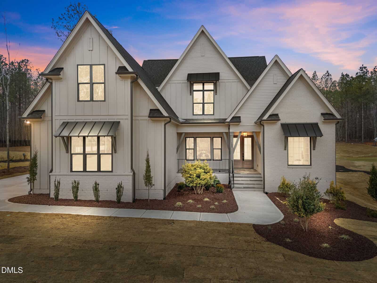 A modern two-story house with light-colored siding and brick, black window trim, multiple gables, covered entryway, landscaped front yard, and a driveway, pictured at dusk with surrounding trees in the background.