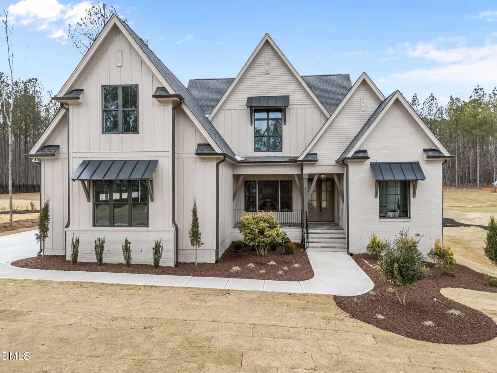 A modern two-story house with light-colored siding and brick exterior, dark window awnings, multiple gables, and a covered front porch, surrounded by minimal landscaping and set against a backdrop of trees.