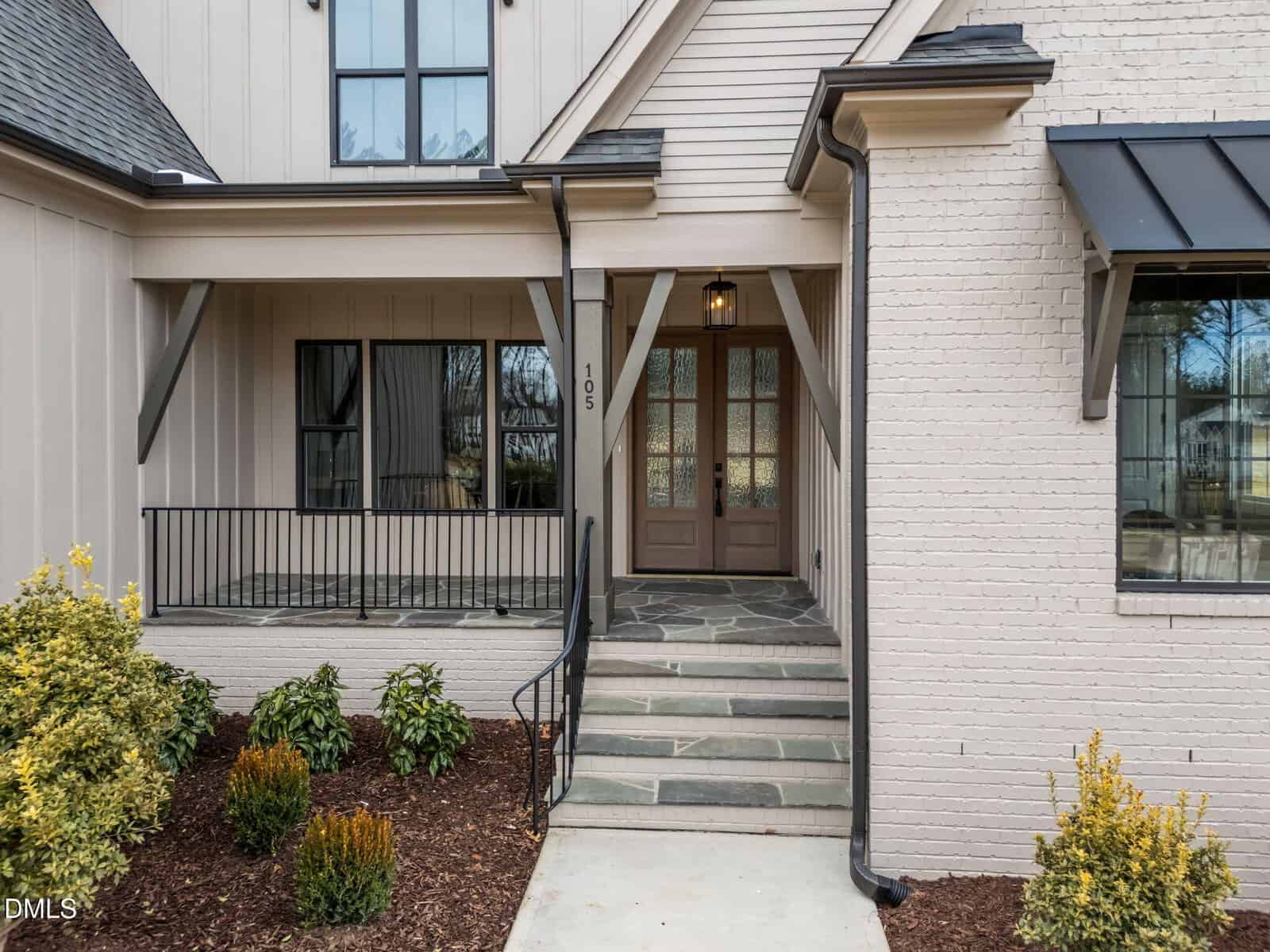 Front entryway of a modern house with light-colored brick and vertical siding, a covered porch, black railing, stone steps, glass-paneled double doors, and landscaped bushes along the walkway.