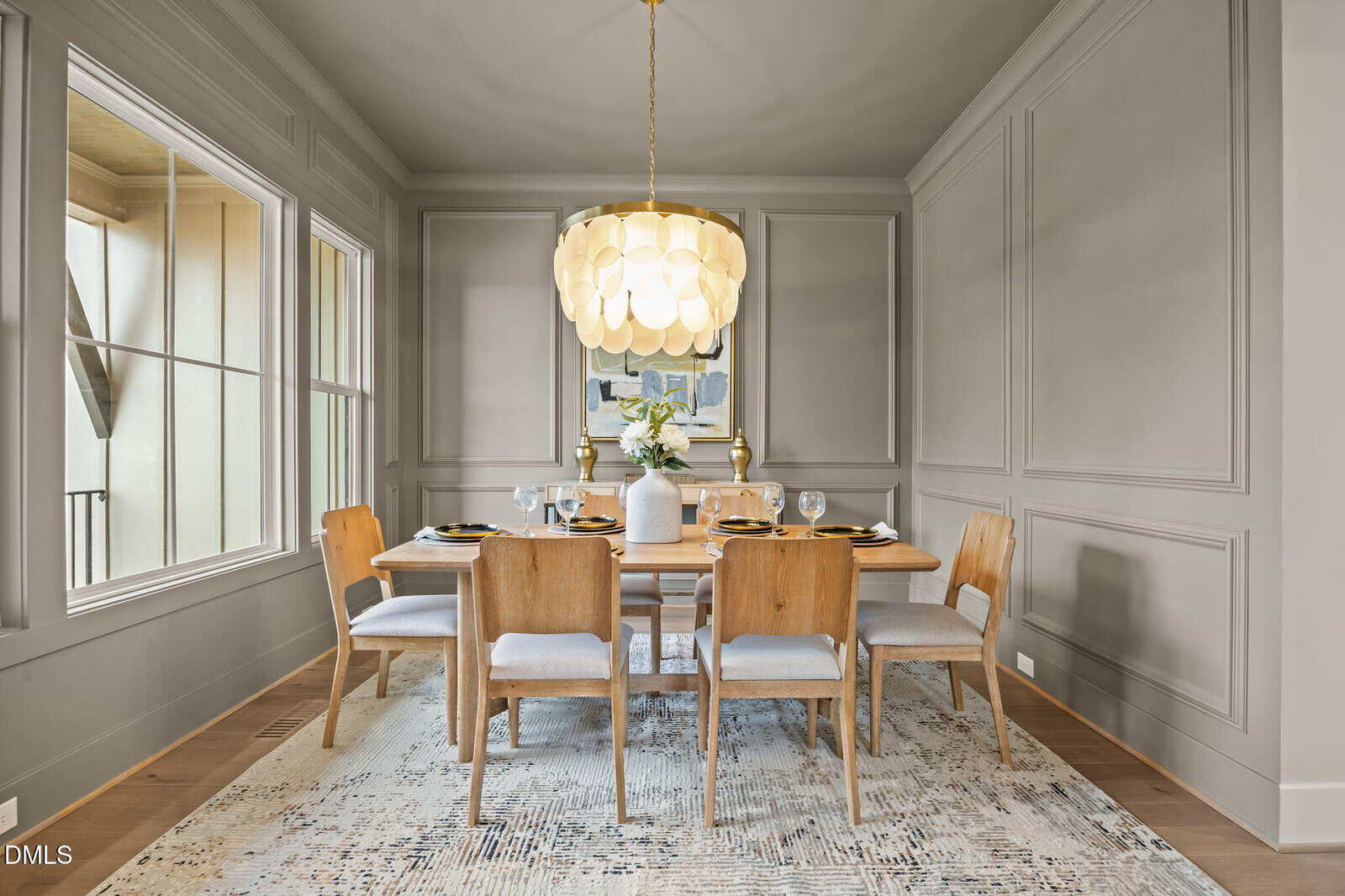 Modern dining room with a wooden table, six chairs, a large round chandelier, and a decorative rug. Neutral-toned walls and a window provide a bright, elegant atmosphere.