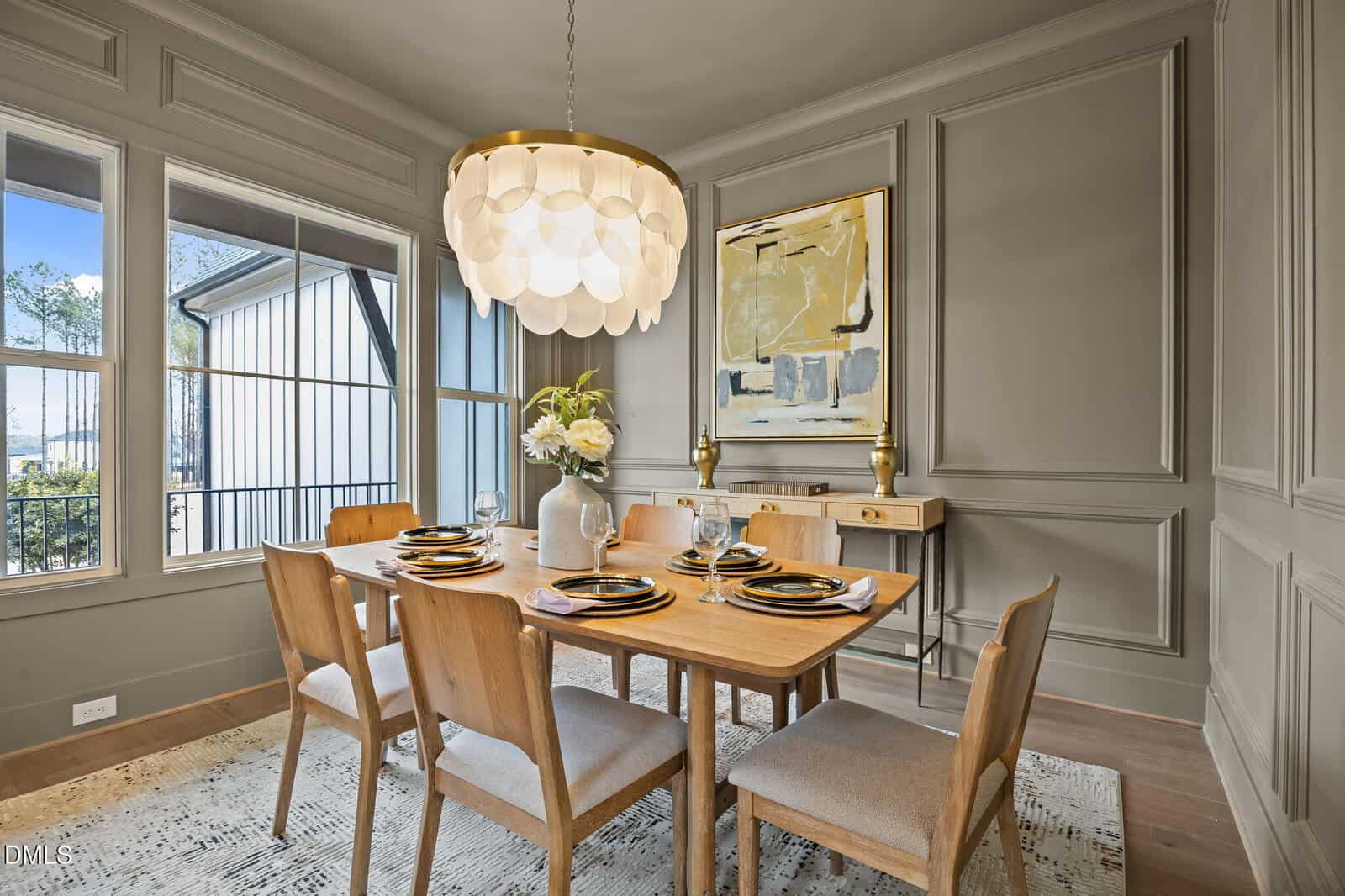 Modern dining room with a wooden table set for six, beige upholstered chairs, a large contemporary chandelier, floral centerpiece, abstract wall art, and a window letting in natural light.