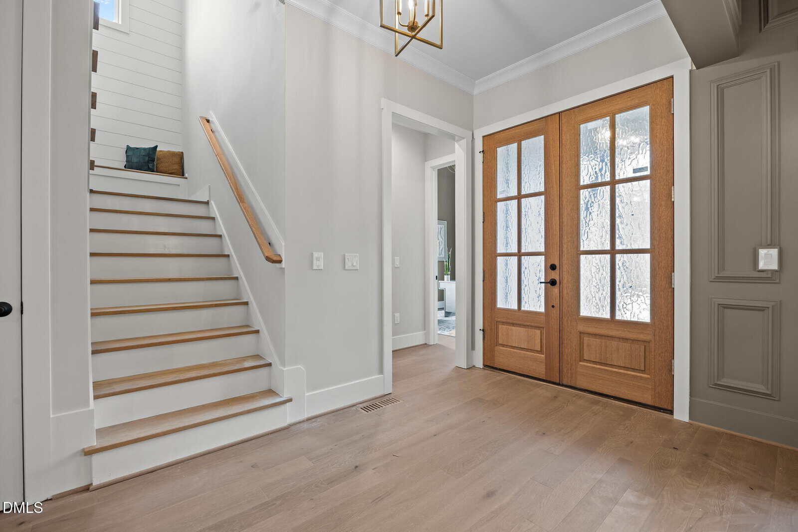 A bright entryway with light wood floors, a staircase with white risers and wood treads, a wooden front door with glass panels, and neutral-colored walls.