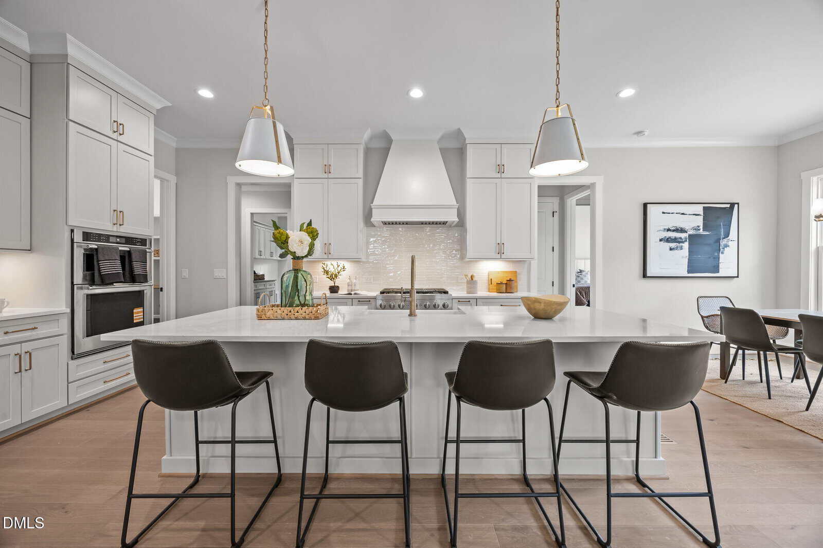 Modern kitchen with white cabinets, stainless steel appliances, a large island with four dark barstools, pendant lights above, and decorative items. Dining area visible in the background.