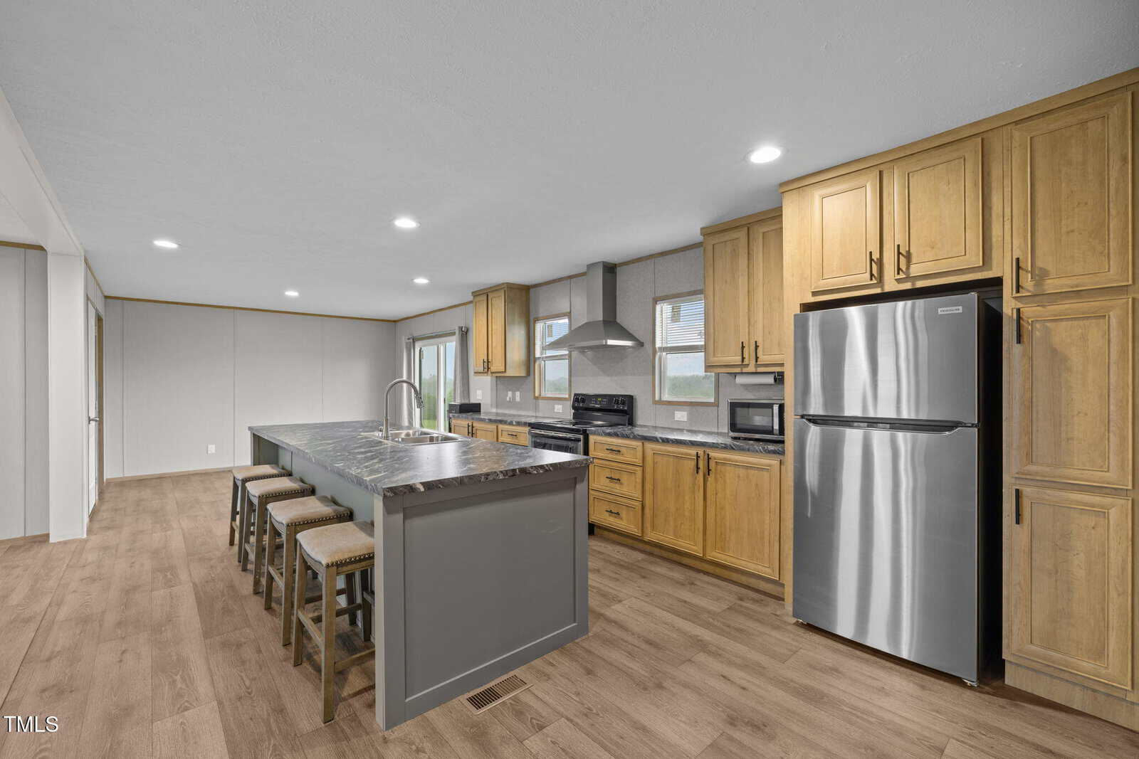 Modern kitchen with wood floors, light wood cabinets, stainless steel appliances, a large gray island with a marble countertop, sink, and three barstools. Ample lighting from ceiling fixtures and windows.